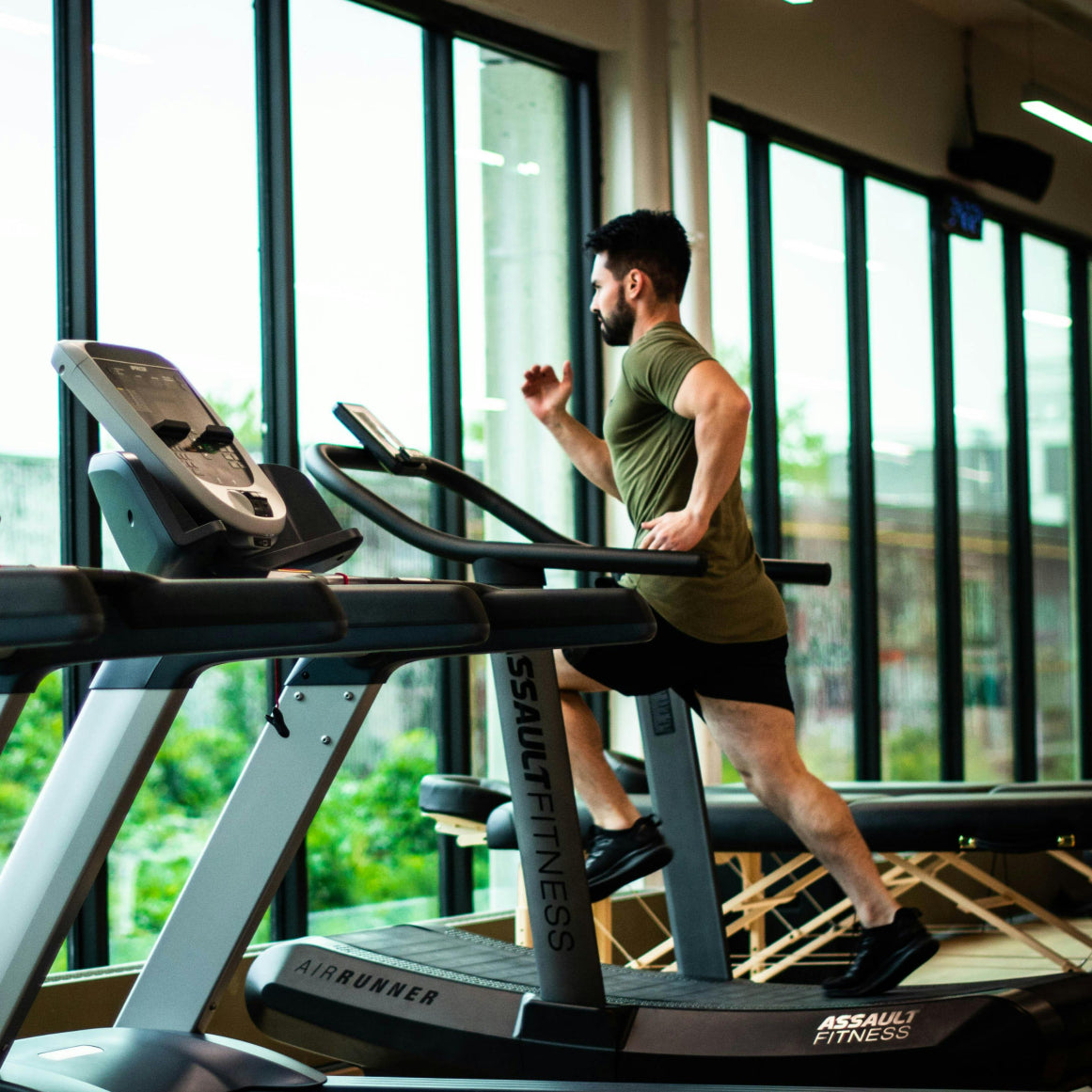 Mand løber på løbebånd i et moderne fitnesscenter med store vinduer. Fokus på fysisk træning, sundhed og en aktiv livsstil.